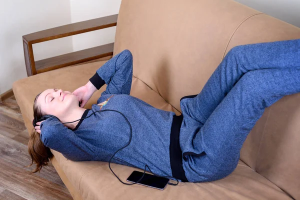 Vacker ung kvinna liggande på soffan med slutna ögon på hörlurar och lyssnar på musik. Från soffan hemma — Stockfoto