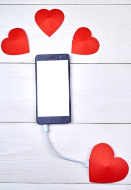 Mobil akıllı telefon ile kopya alanı beyaz ahşap arka plan üzerinde kırmızı kağıt kalp ile şarj beyaz boş perde. Sevgililer günü için mockup şablonu. Aşk, teknoloji kavramı. Üstten görünüm, düz yatıyordu