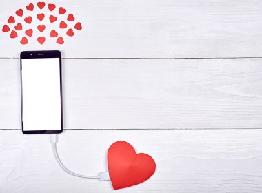 Mobil akıllı telefon ile kopya alanı beyaz ahşap arka plan üzerinde kırmızı kağıt kalp ile şarj beyaz boş perde. Sevgililer günü için mockup şablonu. Aşk, teknoloji kavramı. Üstten görünüm, düz yatıyordu