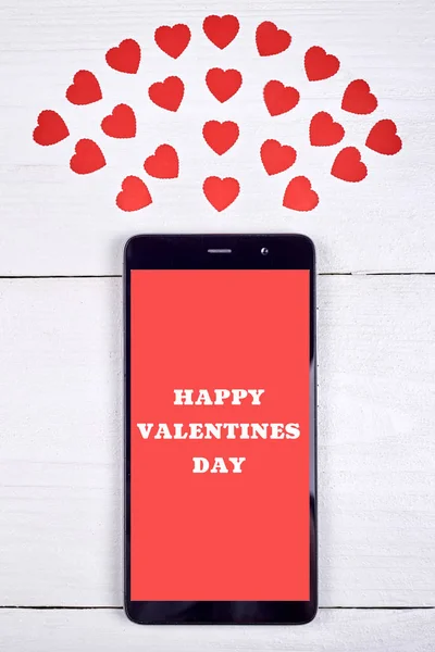Close up of mobile smart phone with sign Happy Valentines Day and red paper hearts on wooden background, copy space. Flat lay, top view. Application mock up template. Love concept