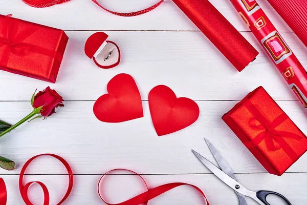 Gift wrapping background. Rolls of red wrapping paper, ribbon, red rose, giamond ring, two hearts, gift boxes and scissors on wooden background, free space. Flat lay, top view