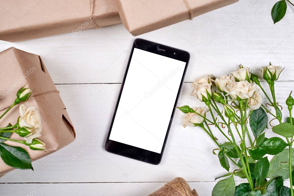 Gift boxes wrapped in kraft with fresh roses and mobile smartphone with white screen on wooden background, copy space. Greeting card mockup. Top view, flat lay. Mobile app presentation