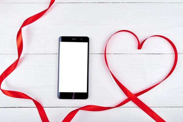 Bovenaanzicht van mobiele smartphone met witte leeg scherm en rood lint gevormd als hart op houten achtergrond, kopie ruimte. Plat lag, top uitzicht. Valentijnsdag toepassingssjabloon mockup — Stockfoto