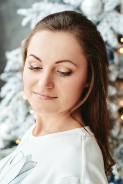 Beyaz süveterli güzel bir kadın Noel ağacına karşı duruyor. Evde takılar ve çelenklerle süslenmiş.