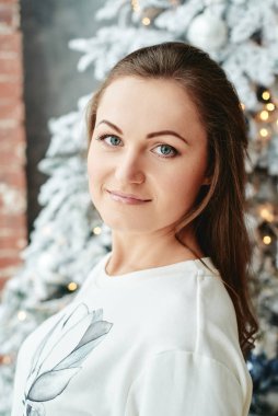 Beyaz kazaklı genç bir kadın Noel ağacına karşı duruyor. Evde takılar ve çelenklerle süslenmiş.