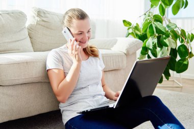 Mutlu gülümseyen genç bir kadının portresi yerde yatıyor, dizüstü bilgisayar kullanıyor ve evde telefonla konuşuyor.