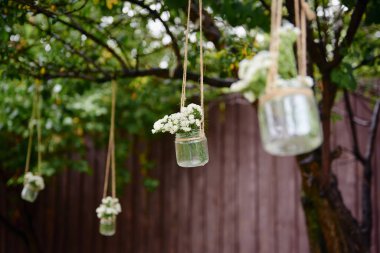 Kırsal düğün süslemeleri, cam kavanozda çiçekler açık havada yeşil ağaçta asılı.