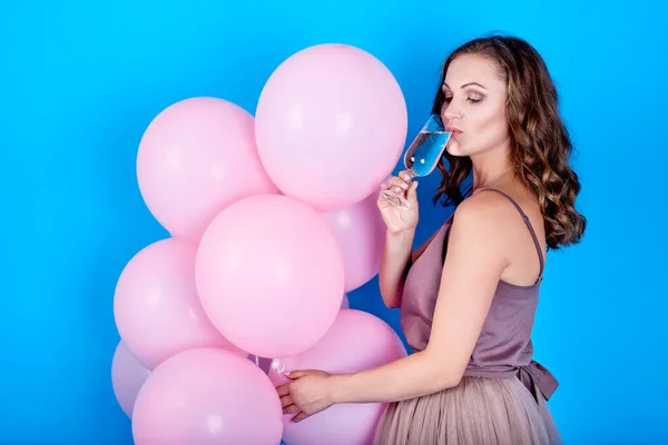 Счастливая молодая женщина в платье с розовыми воздушными шарами и пить шампанское на голубом фоне — стоковое фото