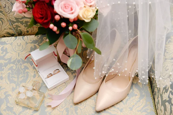 Bouquet da sposa rosa, rose rosse e verde con nastro di raso, scarpe beige da donna, velo e due fedi nuziali dorate su poltrona — Foto Stock