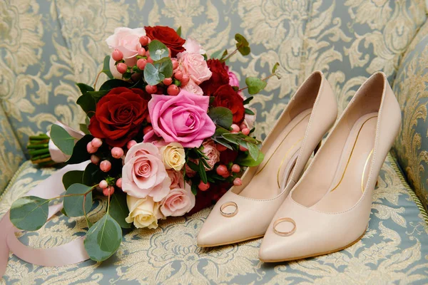 Свадебный букет из розовых, красных роз и зелени с атласной лентой, бежевая женская обувь, вуаль и два золотых обручальных кольца на кресле — стоковое фото