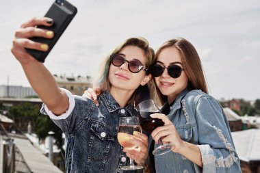 İki genç, gülümseyen kadın bira içiyor, selfie çekiyor ve çatıda eğleniyor.