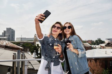 İki genç, gülümseyen kadın bira içiyor, selfie çekiyor ve çatıda eğleniyor.