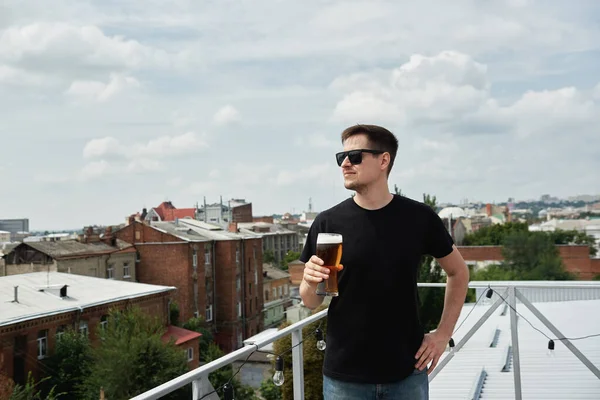 Happy man in black shirt and eyewear drinking beer at bar or pub on roof