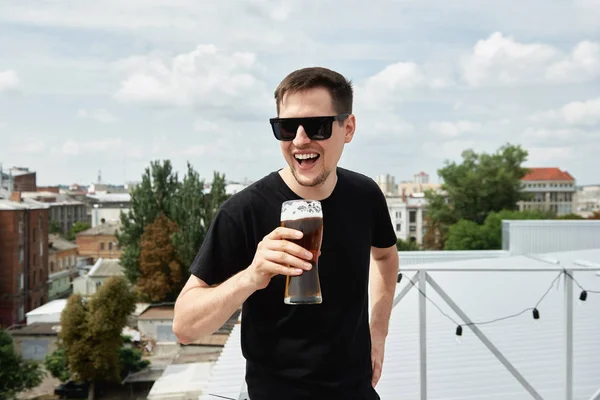 Happy man in black shirt and eyewear drinking beer at bar or pub on roof