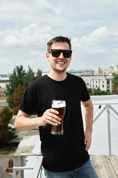 Happy man in black shirt and eyewear drinking beer at bar or pub on roof — 스톡 사진