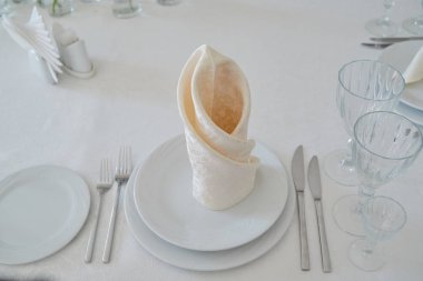 Masada beyaz tabaklar, peçeteler, şarap bardakları, çatal bıçak takımı ve çiçekler var. Düğün resepsiyonunda yer