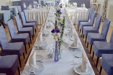 Masada beyaz tabaklar, peçeteler, şarap bardakları, çatal bıçak takımı ve çiçekler var. Düğün resepsiyonunda yer