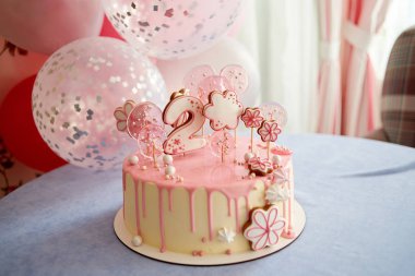 Round pink birthday cake decorated with lollipops, gingerbreads, flowers, beads number two on the top