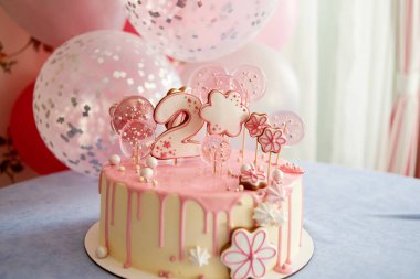 Round pink birthday cake decorated with lollipops, gingerbreads, flowers, beads number two on the top