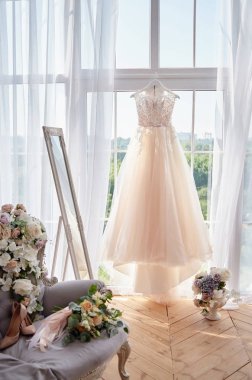 Beautiful beige wedding dress hanging on hanger against window in hotel room