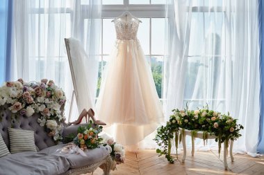 Beautiful beige wedding dress hanging on hanger against window in hotel room
