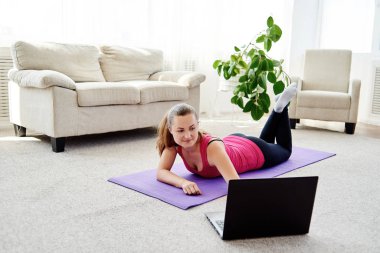 Gülümseyen genç bir kadın evde egzersiz yapıyor ve dizüstü bilgisayar, online eğitim, fotokopi odası kullanıyor. Yoga, pilates egzersizi. Spor, sağlıklı yaşam tarzı konsepti 