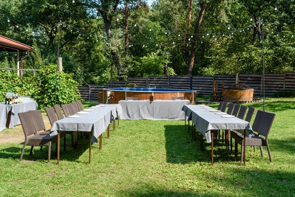 Wedding or birthday table setting with plates, cutlery on table and rattan chairs on green grass outdoors, copy space. Wedding reception, banquet — Stock Photo, Image