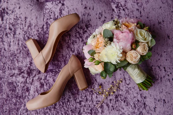 Bridal bouquet of pink peonies, coral roses and greenery with satin ribbon, beige women shoes and hair pins on purple velours armchair, copy space. Wedding concept. Top view, flat lay