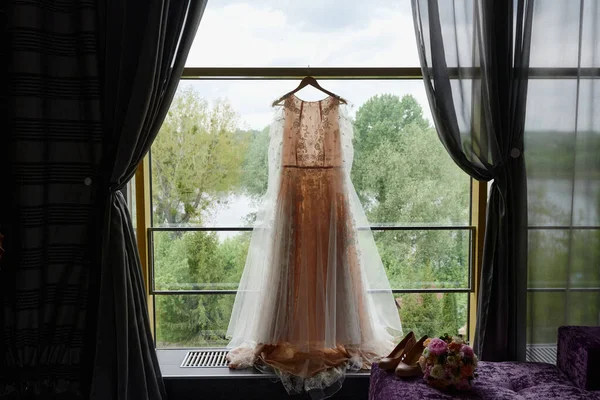 Vacker beige bröllopsklänning hängande på hängare mot fönster i hotellrum, kopiera utrymme. Brudbukett och damskor står på soffan — Stockfoto