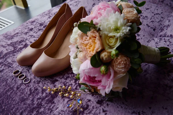 Свадебный букет из розовых пионов, коралловых роз и зелени с атласной лентой, бежевые женские туфли, шпильки и золотые обручальные кольца на фиолетовом велюре кресло, копировальное пространство. Концепция свадьбы — стоковое фото