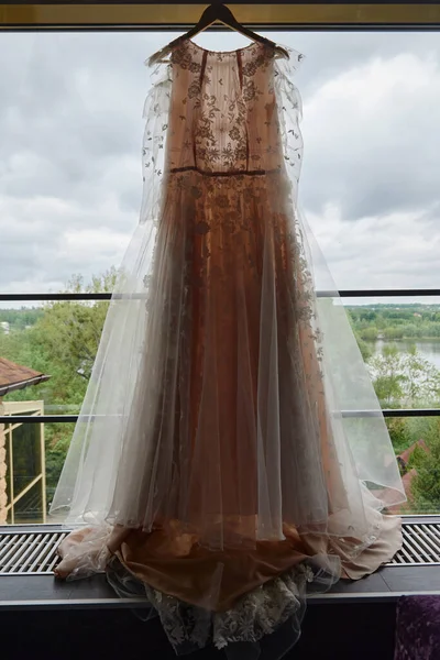 Vestido de noiva bonito bege bordado pendurado no cabide contra a janela na sala, espaço de cópia, de perto. Preparativos da manhã nupcial — Fotografia de Stock