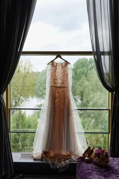 Vacker beige bröllopsklänning hängande på hängare mot fönster i hotellrum, kopiera utrymme. Brudbukett och damskor står på soffan — Stockfoto