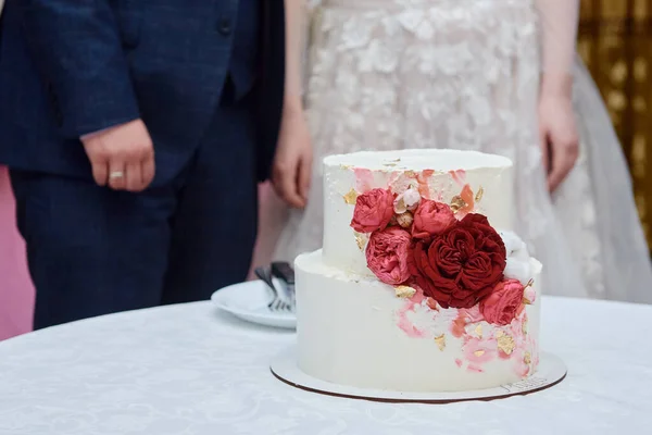 Elegante bolo de casamento branco decorado com rosa e vermelho rosa flores na mesa no restaurante com noiva e noivo no fundo, espaço de cópia — Fotografia de Stock