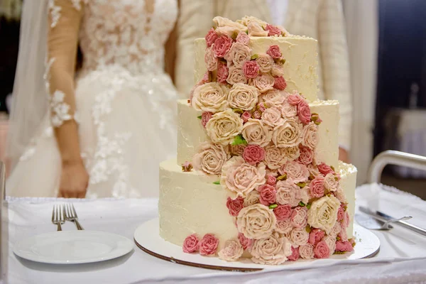 Elegant white wedding cake decorated with pink rose flowers on table in restaurant with bride and groom on background, copy space