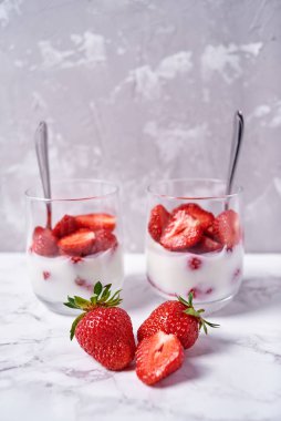 Beyaz masa arkasında taze dilimlenmiş çilekli iki bardak sağlıklı yoğurt, kopyalama alanı. Sağlıklı kahvaltı, yemek konsepti