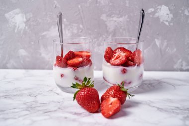 Beyaz masa arkasında taze dilimlenmiş çilekli iki bardak sağlıklı yoğurt, kopyalama alanı. Sağlıklı kahvaltı, yemek konsepti