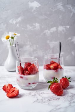 İki bardak sağlıklı yoğurt, taze dilimlenmiş çilek, kaşıklar ve vazoda papatya çiçekleri beyaz masa arkasında, kopyalama alanı. Sağlıklı kahvaltı, yemek konsepti