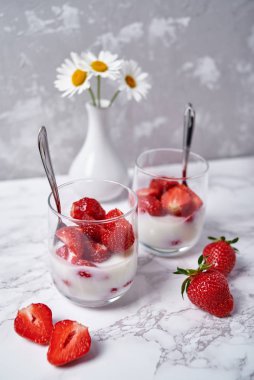 İki bardak sağlıklı yoğurt, taze dilimlenmiş çilek, kaşıklar ve vazoda papatya çiçekleri beyaz masa arkasında, kopyalama alanı. Sağlıklı kahvaltı, yemek konsepti