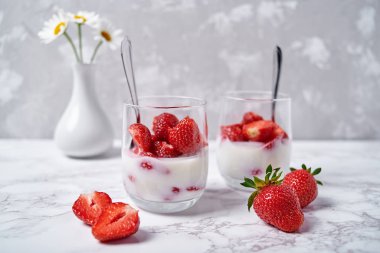 İki bardak sağlıklı yoğurt, taze dilimlenmiş çilek, kaşıklar ve vazoda papatya çiçekleri beyaz masa arkasında, kopyalama alanı. Sağlıklı kahvaltı, yemek konsepti