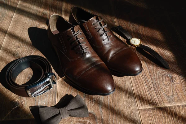 Close up of brown male shoes, bowtie, belt and watches on wood floor background, copy space. Modern man accessories. Wedding details