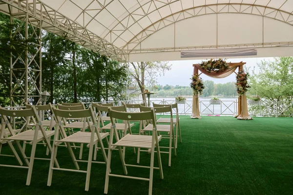 Place for wedding ceremony in tent outdoors, copy space. Wedding arch decorated with flowers and chairs on each side of archway outdoors. Wedding setting