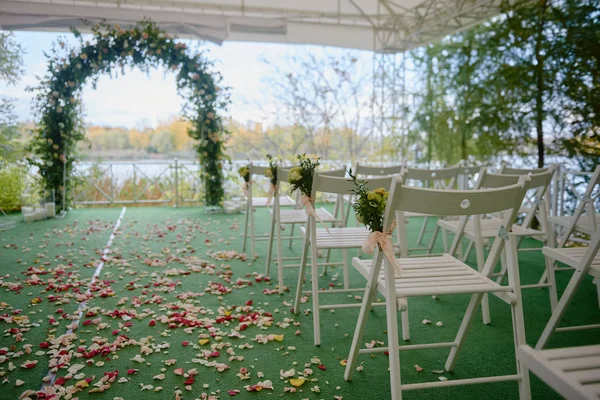 Place for wedding ceremony in tent outdoors, copy space. Wedding arch decorated with flowers and chairs on each side of archway. Wedding setting