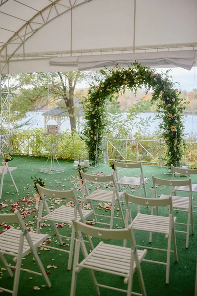 Place for wedding ceremony in tent outdoors, copy space. Wedding arch decorated with flowers and chairs on each side of archway. Wedding setting