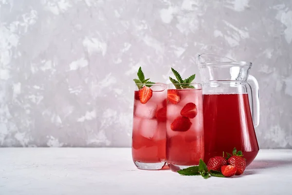 Fresh strawberry lemonade with ice and mint in glasses and jug on white table, copy space. Cold summer drink. Sparkling glass with berry cocktail