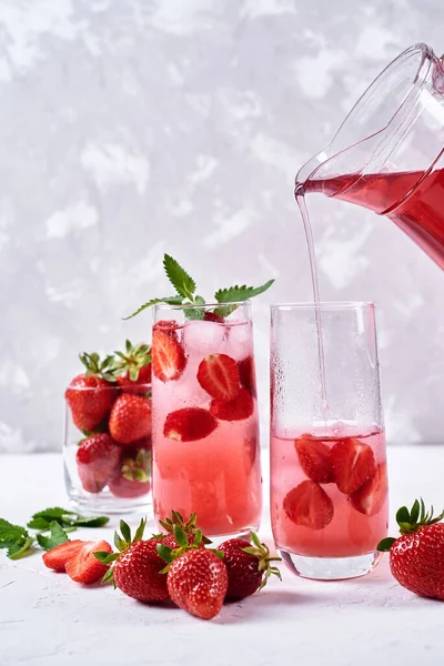 Strawberry lemonade pouring in glass from jug. Cocktail with strawberry, ice and mint in glasses on white concrete background, copy space. Refreshing summer berry drink