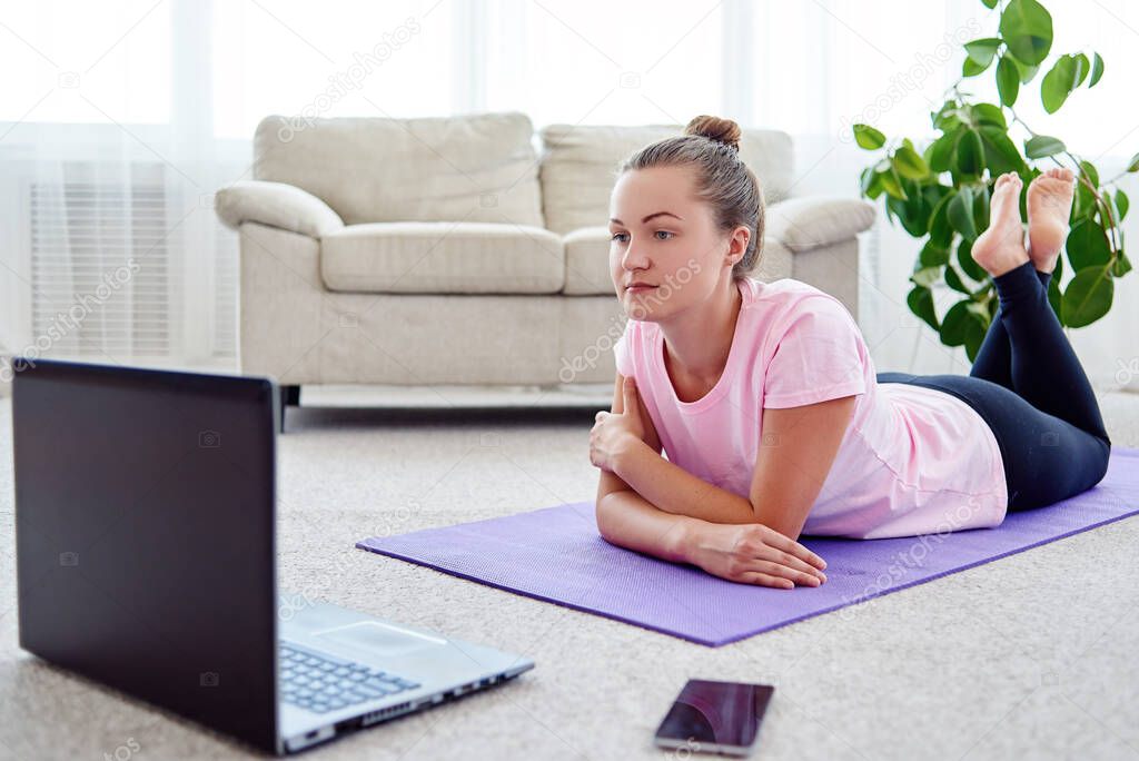Beautiful young woman doing exercise on floor at home, online training on laptop computer, copy space. Full length portrait. Yoga, pilates, working out exercising