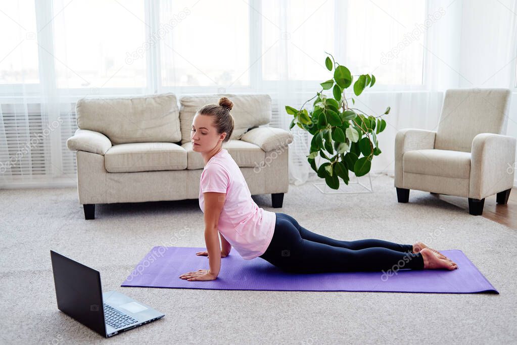 Young woman practicing yoga with laptop at home indoor, copy space. Girl practicing cobra pose, full length. Relaxing and doing yoga. Wellness and healthy lifestyle. Bhujangasana