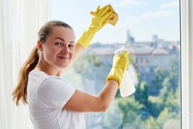 Beyaz gömlekli ve sarı lastik eldivenli bir kadın pencereyi temizleyici sprey ve sarı bir bez parçasıyla temizliyor. İnsanlar, ev işleri ve ev idaresi kavramı