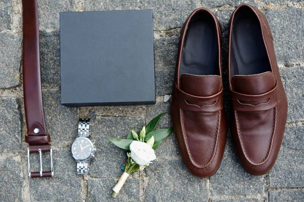 Close up of brown male shoes, belt, watches and boutonniere on stone background, copy space. Modern man accessories. Wedding details