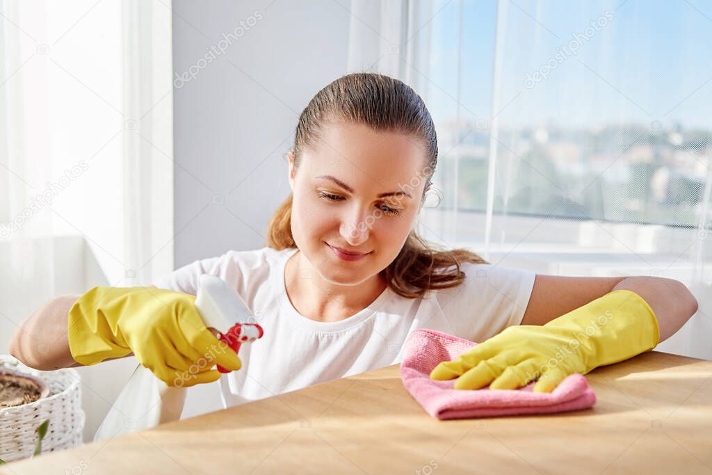 Happy woman in white shirt and yellow protective rubber gloves cleaning at home and wiping dust on wood tablel with sprayer and rag, copy space. Housework, cleanig and chores concept